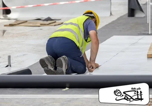 عزل أسطح بالفوم بالفجيرة تُصور هذه اللقطة فنياً يرتدي خوذة وقائية وسترة عاكسة وهو ينكب على الأرض لتركيب أو قياس صفائح عازلة بيضاء اللون