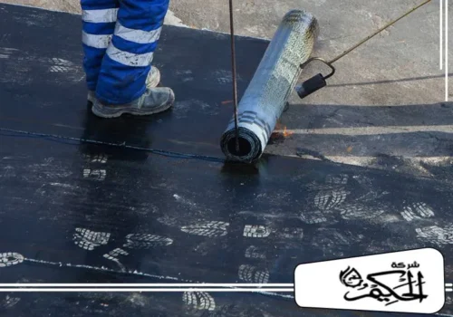 عزل اسطح المباني في ابوظبي عامل بملابس زرقاء يدحرج غشاء بيتومين وتظهر آثار أقدام على السطح.