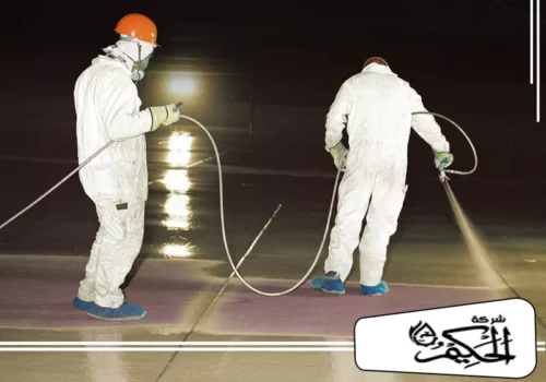عزل خزانات واسطح في دبي 0547842313 شركة الحكيم عاملان يرتديان ملابس واقية كاملة وأقنعة تنفس، وهما يقومان برش طبقة عازلة أو واقية على أرضية خزان كبير.