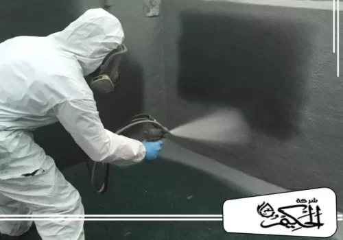 غسيل خزانات مع العزل والتعقيم في دبي 0547842313 شركة الحكيم عامل يرتدي قناع تنفس شامل وبدلة واقية، وهو يقوم برش مادة عازلة أو طلاء على جدار رأسي داخلي للخزان